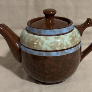 May include: A brown ceramic teapot with a glossy finish, featuring a lid with a knob handle. The body is decorated with a horizontal band of light blue and a mottled pattern of pale green and tan, while the base is a solid dark brown.
