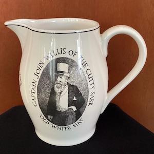 May include: White ceramic pitcher with a curved handle and spout. The pitcher features a black and white illustration of a man in a top hat, with text that reads "CAPTAIN JOHN WILLIS OF THE CUTTY SARK" and "OLD WHITE HAT."