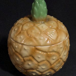 May include: A ceramic pineapple-shaped jar with a green top. The jar has a detailed texture resembling a pineapple.