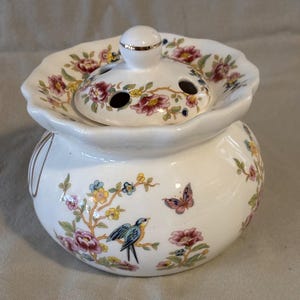 May include: A white ceramic floral arrangement holder with a lid. The lid has three holes and is decorated with flowers and gold trim. The body of the holder features a bird, butterfly, and floral design.