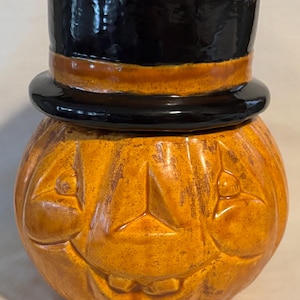 May include: A ceramic Halloween pumpkin with a black top hat lid. The pumpkin is orange with carved facial features, including a smiling mouth and triangular eyes. The hat has a brown band.