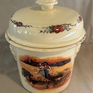May include: A cream-colored enamel canister with a matching lid featuring a floral garland border. The front displays a colorful illustration of two figures in a field. The container has two side handles and a knob on the lid, with visible wear.