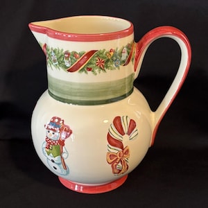 May include: A ceramic Christmas pitcher with a rounded base and a red handle and rim. The pitcher is decorated with a snowman, candy cane, and festive garland. The pitcher is approximately 10 inches tall.