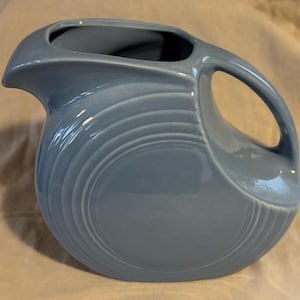 May include: A light blue ceramic pitcher with a curved handle and a spout. The pitcher has a circular design on the front and is approximately 10 inches tall.