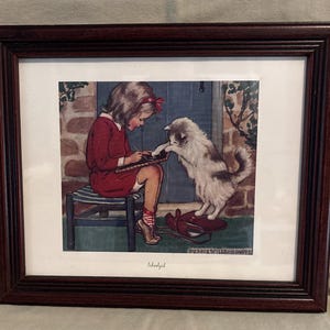 May include: A framed print titled Schoolgirl featuring a child in a red dress sitting on a striped stool writing on a slate while a fluffy white and gray cat stands on a red bag. The artwork is enclosed in a dark brown wooden frame with a white mat.