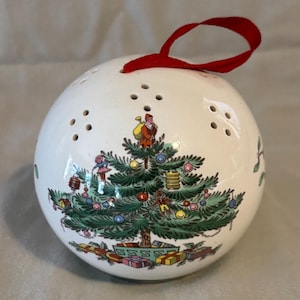 May include: White ceramic Christmas ornament with a red ribbon. The ornament features a detailed illustration of a decorated Christmas tree with Santa Claus and presents. The ornament has small holes for sound.