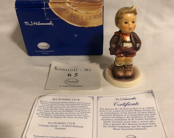 May include: A porcelain figurine of a young boy wearing a brown jacket, red shirt, and blue shorts. The figurine is standing on a small base and has a certificate that reads "M.I. Hummel Certificate".