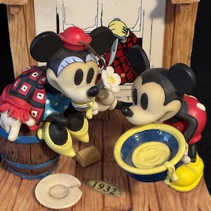 May include: A decorative figurine featuring Mickey and Minnie Mouse in a scene from "The Klondike Kid." Mickey is holding a blue bowl, while Minnie holds a flower. The base includes a barrel, a plate, and the year "1932."