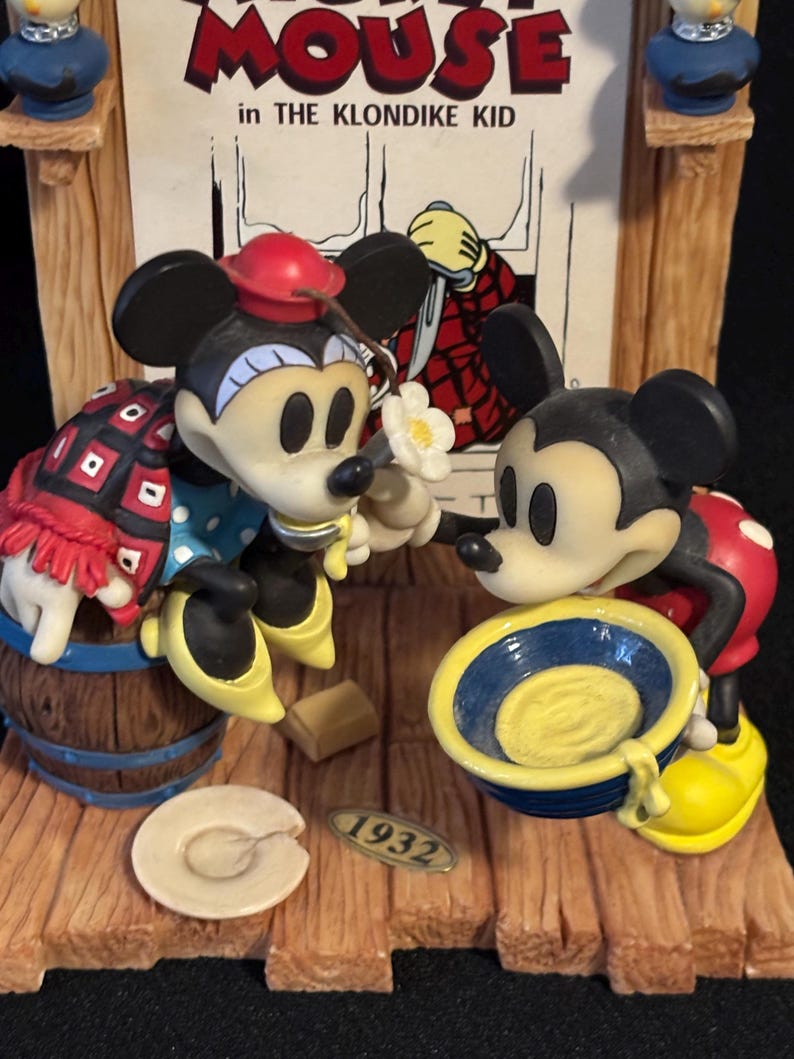 May include: A decorative figurine featuring Mickey and Minnie Mouse in a scene from "The Klondike Kid." Mickey is holding a blue bowl, while Minnie holds a flower. The base includes a barrel, a plate, and the year "1932."