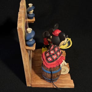 May include: A Disney figurine featuring Mickey Mouse in a red and black patterned shawl, standing near a wooden barrel. The figurine has a red hat and yellow shoes. Two blue vases sit on a wooden shelf. The figurine is set against a dark background.