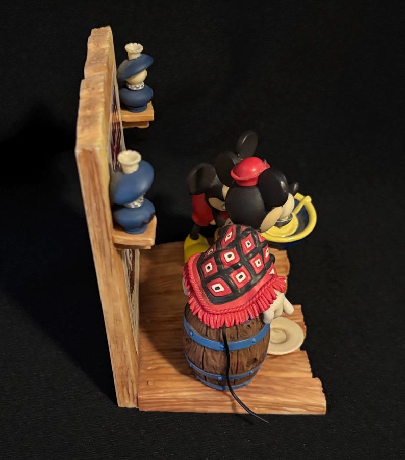 May include: A Disney figurine featuring Mickey Mouse in a red and black patterned shawl, standing near a wooden barrel. The figurine has a red hat and yellow shoes. Two blue vases sit on a wooden shelf. The figurine is set against a dark background.