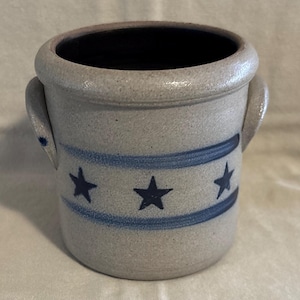 May include: A cylindrical stoneware crock featuring a speckled light gray finish and two side handles. The exterior is decorated with a horizontal band containing three dark blue painted stars, framed by two thin blue lines encircling the vessel.