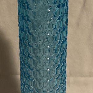 May include: A tall, light blue glass bottle with a textured, bubble-like pattern. The bottle has a rounded top and a slightly flared base. The glass has a glossy finish, reflecting light. The bottle is likely for decorative or functional use.