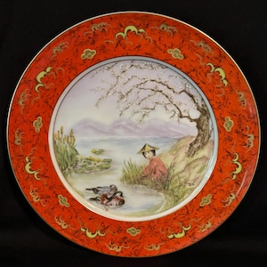 May include: Decorative plate with a vibrant red border featuring gold accents. The central image depicts a serene waterside scene with a figure in traditional attire, ducks, and a blossoming tree. The plate's design evokes a sense of Asian artistry.