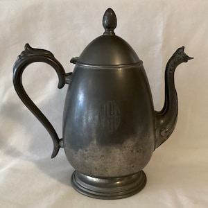 May include: A silver teapot with a rounded body and a decorative handle. The teapot has a lid with a finial and a monogram on the front.