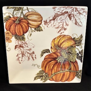 May include: Square ceramic serving dish with a cream-colored background. The dish is decorated with illustrations of orange pumpkins, green leaves, and brown oak leaves, perfect for fall decor.