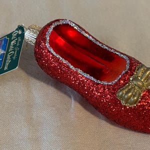 May include: A red glittery glass slipper Christmas ornament with a gold bow. The ornament has a silver trim around the opening. The tag reads "Old World Christmas".