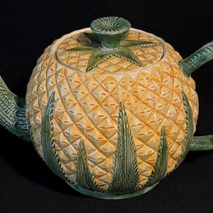 May include: A ceramic teapot shaped like a pineapple. The body is yellow with a textured diamond pattern, and green accents form the handle, spout, lid, and base. The lid has a green star-shaped design. The teapot is on a black background.