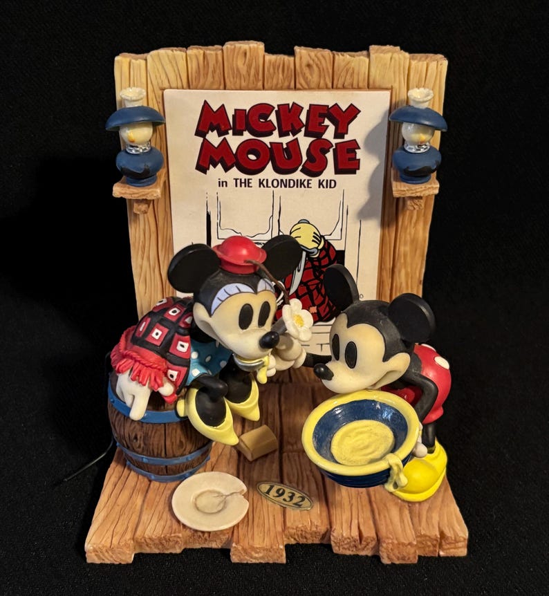 May include: A vintage Mickey Mouse figurine from "The Klondike Kid" featuring Mickey and a friend. The figurine has a wooden frame with two small blue lamps. The year "1932" is printed on the base. Mickey is wearing a red hat and a patterned outfit.