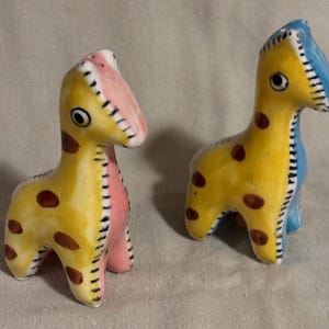 May include: Two ceramic giraffe figurines featuring yellow bodies with brown spots. One giraffe has a pink chest and face detail, while the other has a blue chest and face detail. Both figures have black stitched-style markings along the neck.