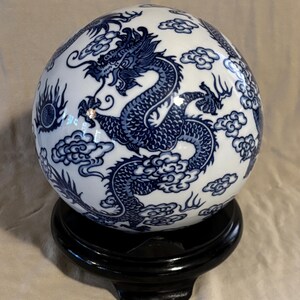 May include: A white ceramic sphere with a blue dragon design and cloud motifs, displayed on a black wooden stand. The dragon is intricately detailed, and the sphere has a glossy finish. A decorative item.