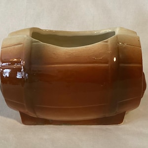 May include: A ceramic barrel-shaped planter with a glossy finish. The planter features a gradient of colors, transitioning from light tan at the top to a rich brown at the base. The design mimics a wooden barrel with horizontal bands.