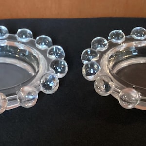 May include: Two clear glass candle holders with a circular design. Each holder has a ring of small, clear glass spheres around the outer edge. The holders sit on a dark surface.