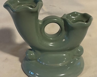 May include: A light green ceramic candle holder with two candle cups and a decorative ring in the center. The holder has a rounded base with three small feet.