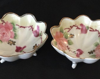 May include: Two white porcelain bowls with three legs each. The bowls are decorated with pink and green floral designs and gold accents.