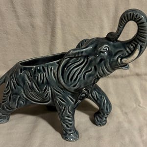 May include: A ceramic elephant planter in a muted teal color. The elephant has detailed carved patterns and a raised trunk. The tusks are white. The planter has a rectangular opening on its back.