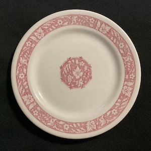 May include: White ceramic plate with a pink floral design around the rim and center. The plate features a repeating pattern of flowers and leaves. The plate is round and has a slightly raised edge.