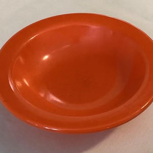 May include: A bright orange, round bowl with a slightly raised rim. The bowl has a smooth, glossy finish and appears to be made of ceramic or a similar material. The bowl is empty and sits on a white surface.