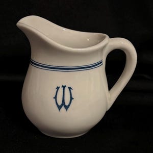 May include: White ceramic pitcher with a curved spout and handle. The pitcher features two horizontal blue stripes near the top and a blue monogram on the front. The pitcher is designed for serving beverages.