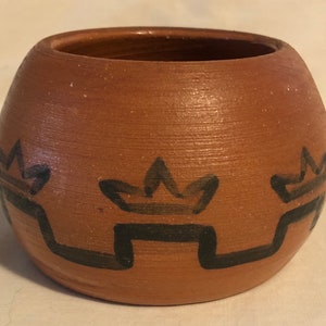May include: A small, round, terracotta pot with a black geometric design. The design features a repeating pattern of stylized crowns and zigzags.
