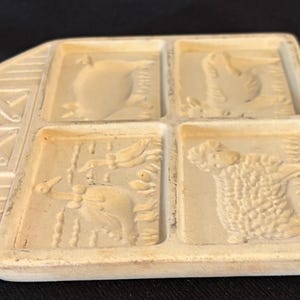 Puede incluir: Un molde de cer&aacute;mica beige y rectangular con cuatro dise&ntilde;os de animales: una manzana, un pollo, un pato y una oveja. El molde tiene un asa decorativa con un dise&ntilde;o en forma de X. Probablemente utilizado para hacer galletas u otros productos horneados.