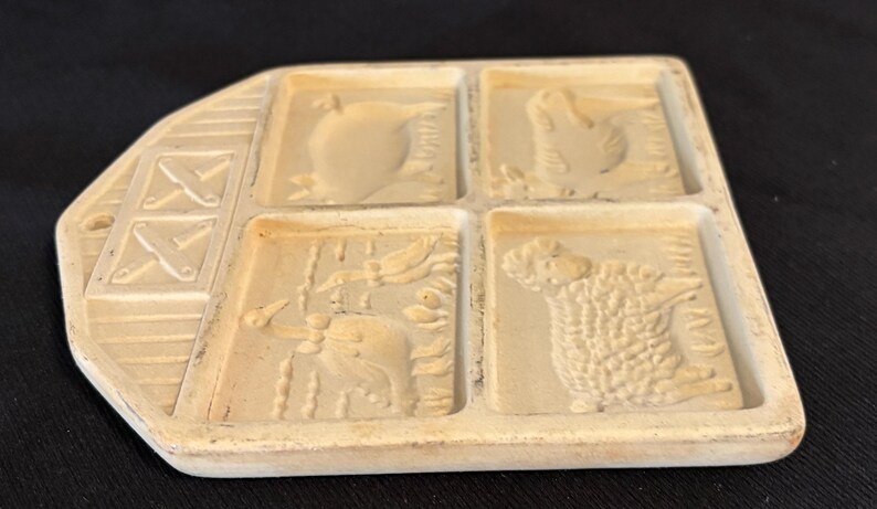 Puede incluir: Un molde de cer&aacute;mica beige y rectangular con cuatro dise&ntilde;os de animales: una manzana, un pollo, un pato y una oveja. El molde tiene un asa decorativa con un dise&ntilde;o en forma de X. Probablemente utilizado para hacer galletas u otros productos horneados.
