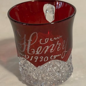 May include: A red glass tumbler with a clear, textured base. The upper portion of the glass is a deep red, with the name "Henry" and the year "1930" etched in white script. The base has a diamond-cut pattern.
