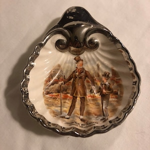 May include: A silver-plated shell-shaped dish with a white porcelain interior. The dish features a hand-painted image of two men in Victorian clothing walking down a street. The text "MICAP. ELE. WT. PRODUCES DAVID. TO. LONDON" is printed on the bottom of the dish.