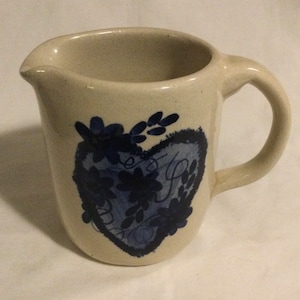 May include: A cream-colored ceramic pitcher with a blue heart design. The heart is surrounded by blue flowers and leaves. The pitcher has a handle and a spout.