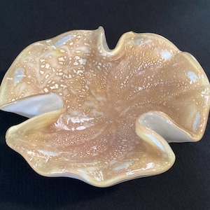 May include: A decorative, free-form glass bowl with a unique shape. The bowl features a blend of tan, cream, and white colors with a speckled pattern. The edges are gently curved, creating an organic, artistic design.
