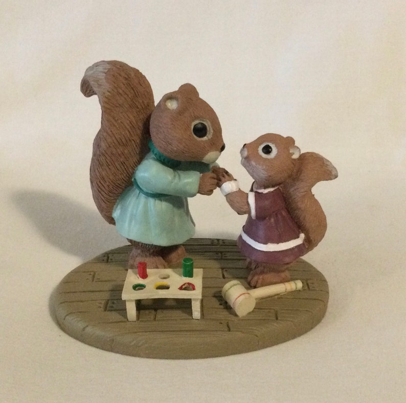 Puede incluir: Una figurita de dos ardillas, una con un vestido azul y la otra con un vestido morado, jugando con un juguete de madera. Las ardillas est&aacute;n de pie sobre una base de madera con un patr&oacute;n de ladrillo.