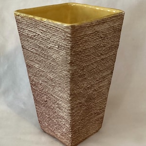 May include: A textured, square-shaped vase with a light brown exterior and a yellow interior. The vase has a slightly tapered design, wider at the top and narrower at the base. The surface has a rough, woven appearance.