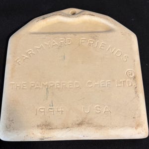 Puede incluir: Un utensilio de cocina Farmyard Friends de cer&aacute;mica color crema, con un agujero para colgar. El texto "THE PAMPERED CHEF LTD. 1994 USA" est&aacute; grabado en relieve en la parte delantera. La herramienta tiene una base redondeada y una parte superior en forma de asa.