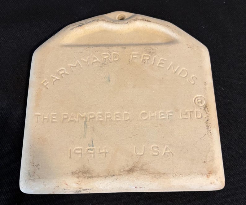 Puede incluir: Un utensilio de cocina Farmyard Friends de cer&aacute;mica color crema, con un agujero para colgar. El texto "THE PAMPERED CHEF LTD. 1994 USA" est&aacute; grabado en relieve en la parte delantera. La herramienta tiene una base redondeada y una parte superior en forma de asa.