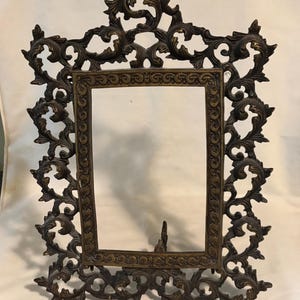 May include: Ornate bronze-colored picture frame with a rectangular opening. The frame features intricate, scrolling leaf and vine designs. A small stand is attached to the back. The frame is designed to hold a photograph or artwork.