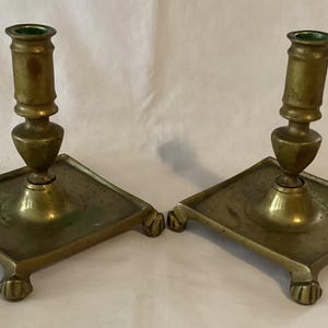 May include: A pair of antique brass candlesticks with square bases and four feet. The candlesticks have a simple design with a round, tapered candle holder.