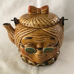 May include: A brown ceramic teapot shaped like a woman's head with a brown and white braided hair, wearing glasses and a brown hat with a pink bow.