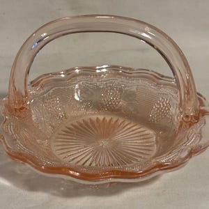 May include: A pink glass candy dish with a scalloped edge and a handle. The dish has a floral design and a textured bottom.