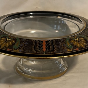 May include: An oval-shaped glass bowl with a decorative rim. The rim is black with gold trim and features a floral design in shades of green, yellow, and orange. The bowl sits on a clear, pedestal base with gold accents.