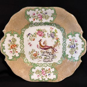 May include: A decorative, scalloped-edge serving platter with a detailed bird and floral design. The center features a bird perched on a rock, surrounded by colorful flowers. The border has a gold-colored, textured pattern with green and white floral accents.